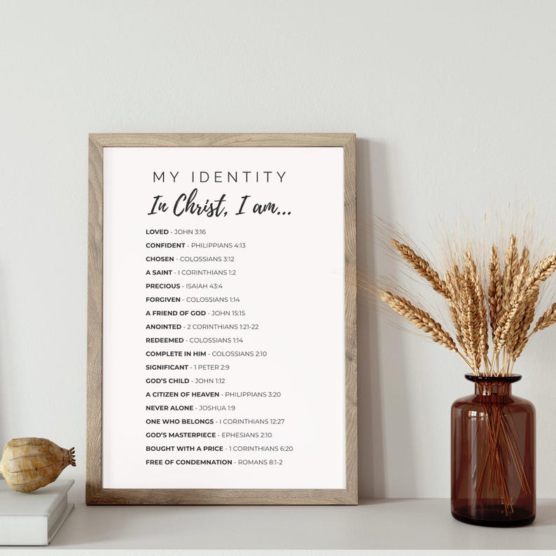 May include: A framed print with the text "MY IDENTITY In Christ, I am..." and a list of affirmations. The frame is light brown. A brown glass vase with wheat stalks is to the right. A small, decorative object sits on a white surface.