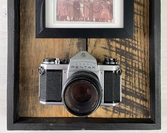 May include: A vintage silver Asahi Pentax camera with a black leather strap and a black lens with the text "SMC Takumar 1:1.8/55 Asahi Opt. Co., Japan" is displayed in a wooden frame with a black mat and a black and white photo of a group of people.