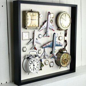 May include: A black shadow box frame containing a collection of vintage-style analog clocks, wristwatches, and die-cast model airplanes arranged on a light-colored fabric backing. The display includes various round and square timepieces.