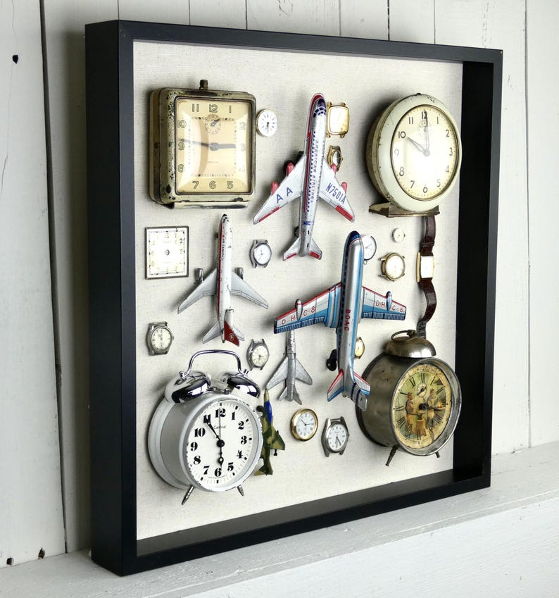 May include: A black shadow box frame containing a collection of vintage-style analog clocks, wristwatches, and die-cast model airplanes arranged on a light-colored fabric backing. The display includes various round and square timepieces.
