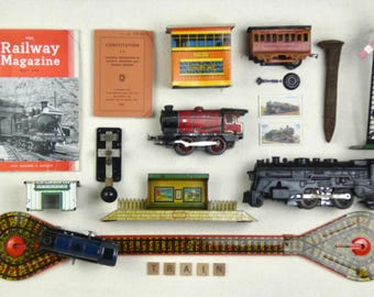 May include: A collection of vintage toy train items including The Railway Magazine, a Constitution booklet, a tin station, a red and black locomotive, a black steam engine, a signal post, a metal spike, and a long track with circular ends.