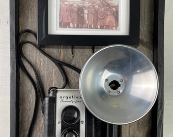 May include: A vintage black and silver argoflex seventy-five camera with a flash attachment, a black strap, and a black frame with a sepia-toned photo of a group of people.
