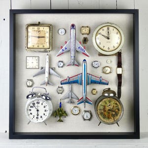 May include: A square black shadow box frame containing a collection of vintage analog clocks, wristwatches, and die-cast model airplanes arranged on a light beige fabric background. The display includes various round and square timepieces.