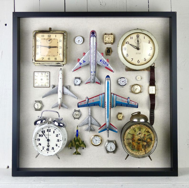 May include: A square black shadow box frame containing a collection of vintage analog clocks, wristwatches, and die-cast model airplanes arranged on a light beige fabric background. The display includes various round and square timepieces.