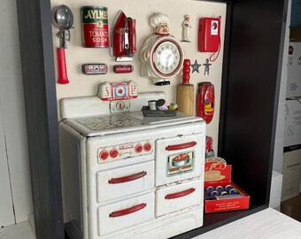 May include: A black shadow box display containing a vintage white toy stove with red accents, surrounded by miniature kitchen collectibles including a tomato soup can, a chef clock, a red iron, a rolling pin, and a red box labeled Cake Decorator.
