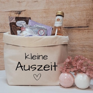 Könnte beinhalten: Eine beigefarbene Canvas-Tragetasche mit den Worten "kleine Auszeit" und einem Herzsymbol auf der Vorderseite. Die Tasche ist mit verschiedenen Artikeln gefüllt, darunter eine Flasche Roséwein und verpackte Leckereien. Zwei Badekugeln und Trockenblumen befinden sich neben der Tasche.