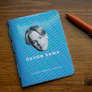 May include: A notebook with a light blue cover featuring the name "DEVON SAWA" and a photo of a person. The cover has repeating text of "DEVON SAWA". An orange pen rests beside the notebook on a wooden surface.