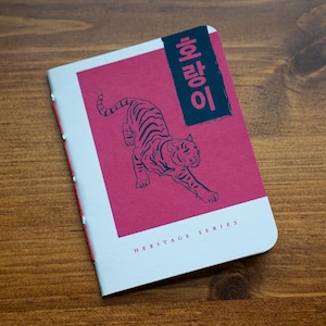 May include: A notebook with a pink cover featuring a tiger illustration and text in Korean. The notebook has a white border and the words "HERITAGE SERIES" are printed at the bottom. The notebook is on a wooden surface.