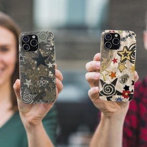 May include: Two phone cases with star designs are held up in front of a blurred background. One case has a dark background with white and gray stars. The other case has a cream background with gold, red, and black stars, and a small disco ball.