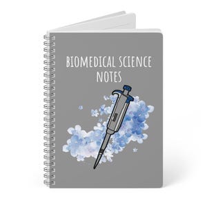 May include: A spiral-bound notebook with a gray cover featuring the text "BIOMEDICAL SCIENCE NOTES." The cover also displays a drawing of a pipette and blue floral accents. The notebook is open, revealing blank pages.
