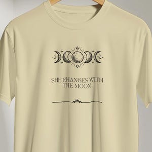 Pode incluir: Uma t-shirt de manga curta em bege claro com um gráfico preto das fases da lua na parte superior. Abaixo das fases lunares, o texto "SHE CHANGES WITH THE MOON" está escrito numa fonte serifada, seguido por uma linha horizontal fina central.