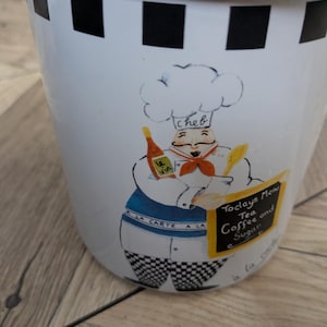 May include: A white ceramic canister featuring a black and white checkered border at the top. The front displays a cartoon chef illustration holding a bottle labelled le vin and a yellow sign with the text Todays Menu Tea Coffee and Sugar.