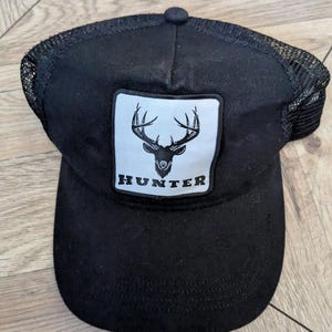 May include: A black trucker cap featuring a white square patch on the front. The patch displays a black graphic of a deer head with antlers above the word HUNTER in bold, black, capital letters. The cap has a solid front panel and mesh back.