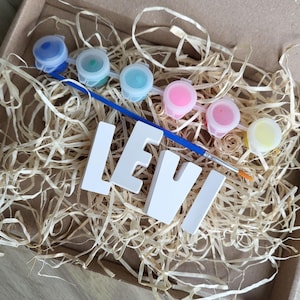 Könnte beinhalten: Ein Bastelset in einem Karton, das Farbtöpfe in verschiedenen Farben, einen Pinsel und den Namen "LEVI" in weißen Großbuchstaben enthält. Die Box ist mit strohähnlichem Verpackungsmaterial gefüllt.