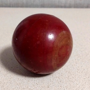 Puede incluir: Un objeto esférico con una superficie lisa de color rojo intenso que presenta una costura visible alrededor de su centro y una mancha circular de color marrón anaranjado más claro en un lado. El objeto tiene un acabado brillante.