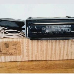 May include: A black vintage car radio with a horizontal frequency display, multiple push buttons, and a separate black speaker connected by a long white cord. The unit rests on a rectangular cardboard box featuring printed shipping icons.