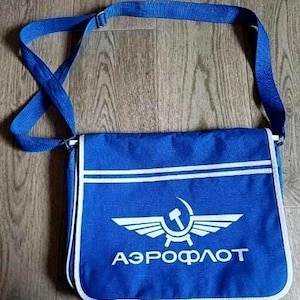 Puede incluir: Un bolso de mensajero azul con correa ajustable y ribetes blancos. La solapa frontal presenta un logotipo gráfico blanco de una hoz y un martillo con alas, acompañado del texto cirílico AEROFLOT impreso en letras blancas.