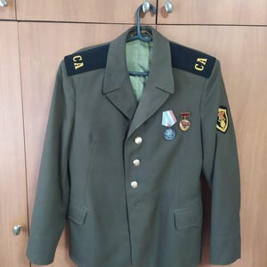 May include: An olive green military-style jacket featuring black shoulder boards with the yellow Cyrillic letters CA, gold-toned buttons, two medals pinned to the chest, and a patch on the left sleeve displaying a red star and a golden emblem.