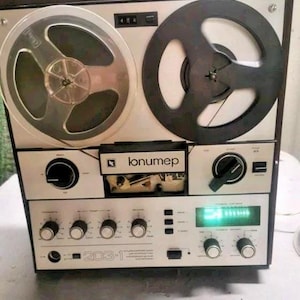 Può includere: Un registratore a bobine vintage con frontalino argentato, due grandi bobine circolari e un pannello di controllo centrale con le scritte "lonumep" e "203-1". Il dispositivo presenta diverse manopole nere, un contatore digitale e un display verde.