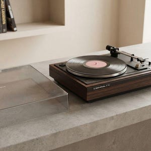 May include: A dark wood grain turntable with a black top surface, a silver platter, and a black tonearm. A black vinyl record with a pink center label is on the platter. A clear, rectangular plastic dust cover sits to the left of the turntable.