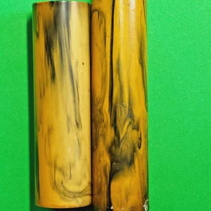 Könnte beinhalten: Zwei zylindrische Objekte mit einem marmorierten Muster aus senfgelben und schwarzen Streifen. Die Oberflächen sind glatt und glänzend und weisen ein durchgehendes, wirbelndes Design auf, das sich um die Länge jedes Zylinders zieht.
