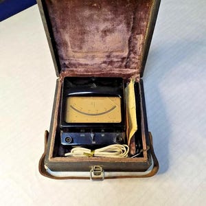 May include: A vintage black light meter housed in a dark rectangular case with a purple velvet interior. The device features a gold-toned analog display with a needle, a central adjustment dial, and a coiled white cord tucked into the bottom compartment.