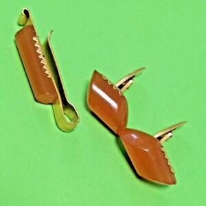 Può includere: Due clip in metallo dorato con inserti in resina liscia color ambra di forma rettangolare. Una clip è un pezzo unico, mentre l'altra è composta da due segmenti in resina color ambra collegati su un supporto in metallo dorato.