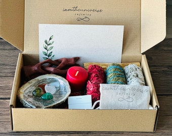 May include: A cardboard box with a variety of items inside, including a red candle, a small wooden sculpture, a shell with crystals, and several bundles of sage. The box is labeled "iamtheuniverse infinity".