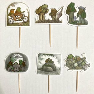 May include: Six cupcake toppers featuring illustrations of two frogs in various activities. The illustrations are in a vintage style with brown, green, and white colors. The frogs are riding a bicycle, sailing a boat, having tea, sitting on a rock, and riding a sled.