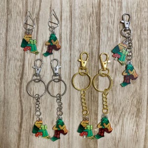 May include: Five keychains featuring a green frog reading a book. Two keychains have a silver chain and a silver ring. Two keychains have a gold chain and a gold ring. One keychain has a silver ring and a silver chain with a frog charm.