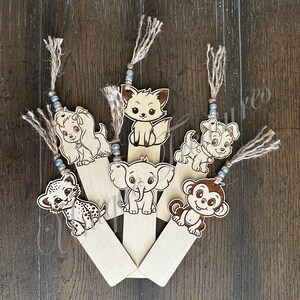 May include: A collection of six wooden bookmarks featuring various animal designs. Each bookmark has a different animal, including a puppy, fox, elephant, monkey, and leopard. The bookmarks are attached to a jute string with beads.