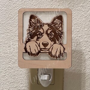 May include: A light-colored wooden night light featuring a laser-engraved illustration of a dog with pointed ears and paws resting on a horizontal ledge. The rectangular wooden frame surrounds the dog portrait, which is mounted on a white base.