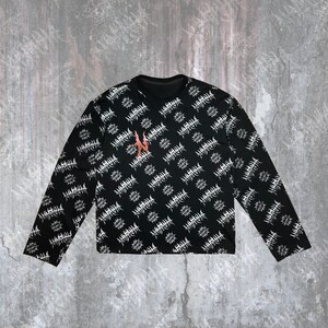 May include: Black long-sleeve shirt with a repeating white graphic pattern. The shirt features a crew neck and a small orange design on the chest. The graphic pattern appears to be a stylized, abstract design.