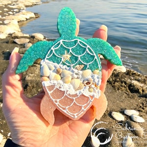 May include: A handcrafted sea turtle figurine with a teal shell, adorned with seashells and a small starfish. The turtle's body is a mix of peach and white resin, held against a beach backdrop.