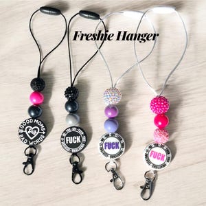 May include: Four beaded lanyards with black, pink, and silver beads. Each lanyard has a round charm with text. The text on the charms includes "GOOD MOMS SAY BAD WORDS" and "FUCK". The lanyards have a black clasp and a black or white cord.