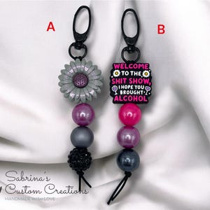 May include: Two keychains with black clasps. One features a gray flower and beads in shades of purple and gray. The other has a charm that reads "WELCOME TO THE SHIT SHOW, I HOPE YOU BROUGHT ALCOHOL" with pink and purple beads.