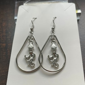 May include: A pair of silver-toned teardrop earrings. Each earring features a silver charm with a stylized candle design. The earrings hang from a silver fish hook.