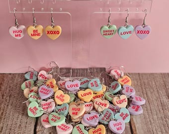 May include: Heart-shaped earrings in pastel shades, inscribed with phrases such as "HUG ME" and "BE MINE." The earrings are displayed on a clear stand and scattered on a wooden surface, evoking a romantic theme.
