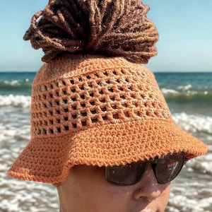 May include: A crocheted orange bucket hat with a wide brim and open weave design. The hat has a brown, textured topknot. The person in the image is wearing sunglasses and a black shirt, with the ocean in the background.