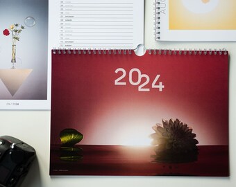 Peut inclure: Un calendrier rouge avec l'année 2024 imprimée en blanc. Le calendrier a une photo d'un coucher de soleil sur une étendue d'eau avec deux plantes de chaque côté du soleil.