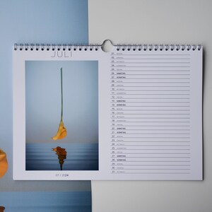 Könnte beinhalten: Ein Wandkalender mit Spiralbindung, der ein Foto einer gelben Calla vor blauem Hintergrund zeigt. Das Wort "JULI" ist oben aufgedruckt, mit dem Datum "07/2024" unten. Die Kalenderseiten haben linierte Felder für tägliche Notizen.