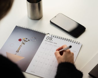 May include: A white calendar with a black spiral binding. The calendar is open to September 2024, with the 9th circled. A person's hand is writing on the calendar with a white pen.