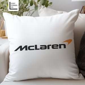 Könnte beinhalten: Ein weißes Kissen mit einem schwarzen McLaren-Logo und einem orangefarbenen Akzent. Das Logo ist ein stilisiertes Wortzeichen mit einer geschwungenen Linie.