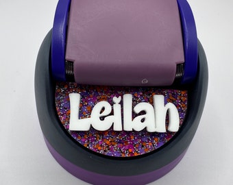 Puede incluir: Troquel artesanal morado y negro con una base de purpurina blanca y el nombre "Leilah" en letras blancas.