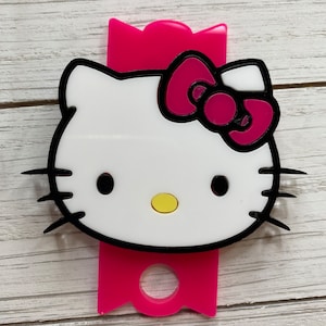 May include: A pink Hello Kitty keyring with a black outline and a white face. The keyring has a black bow with a pink bow on top.