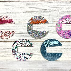 May include: Five round, colorful, glitter-filled, resin nameplate holders with white lettering. The names are "Julianna", "Julianna", "Julianna", "Isela", and "Avanna".