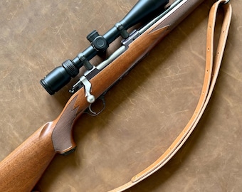 Peut inclure: Une carabine à verrou dotée d'une crosse en bois poli avec des zones de préhension texturées, un canon en métal noir mat et une lunette de visée noire montée. Une sangle en cuir beige clair est fixée à l'arme par des attaches métalliques.