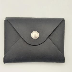May include: A black envelope-style card holder featuring a smooth, matte finish and a single silver-toned metal snap button closure centered on the triangular flap. The rectangular accessory has clean, folded edges and a minimalist, flat design.