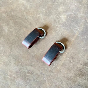 May include: Two dark brown leather loops, each folded and secured with a silver-toned metal D-ring. The leather has a smooth, slightly distressed finish with visible grain and darker shading along the edges of the folded material.