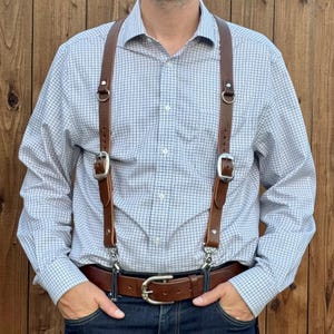 May include: A pair of brown leather suspenders with silver-toned metal buckles and hardware, worn over a light blue and white grid-patterned long-sleeve button-down shirt and a brown leather belt with a silver-toned metal buckle.